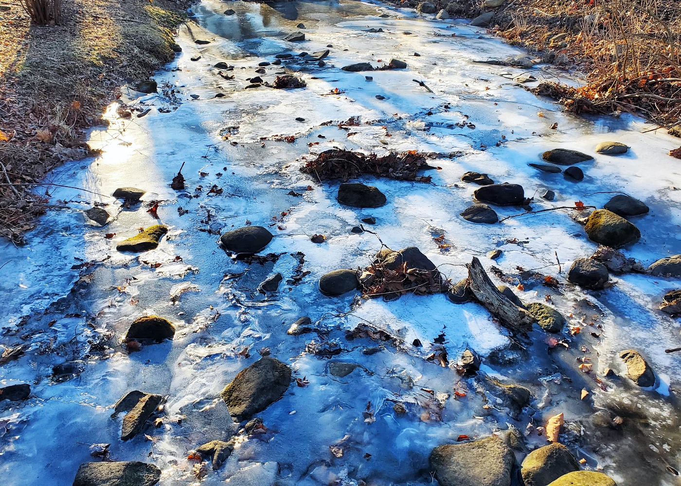 A frozen stream in winter during the golden hour, with sunlight making it shimmer and shadows of trees painting it orange and blue