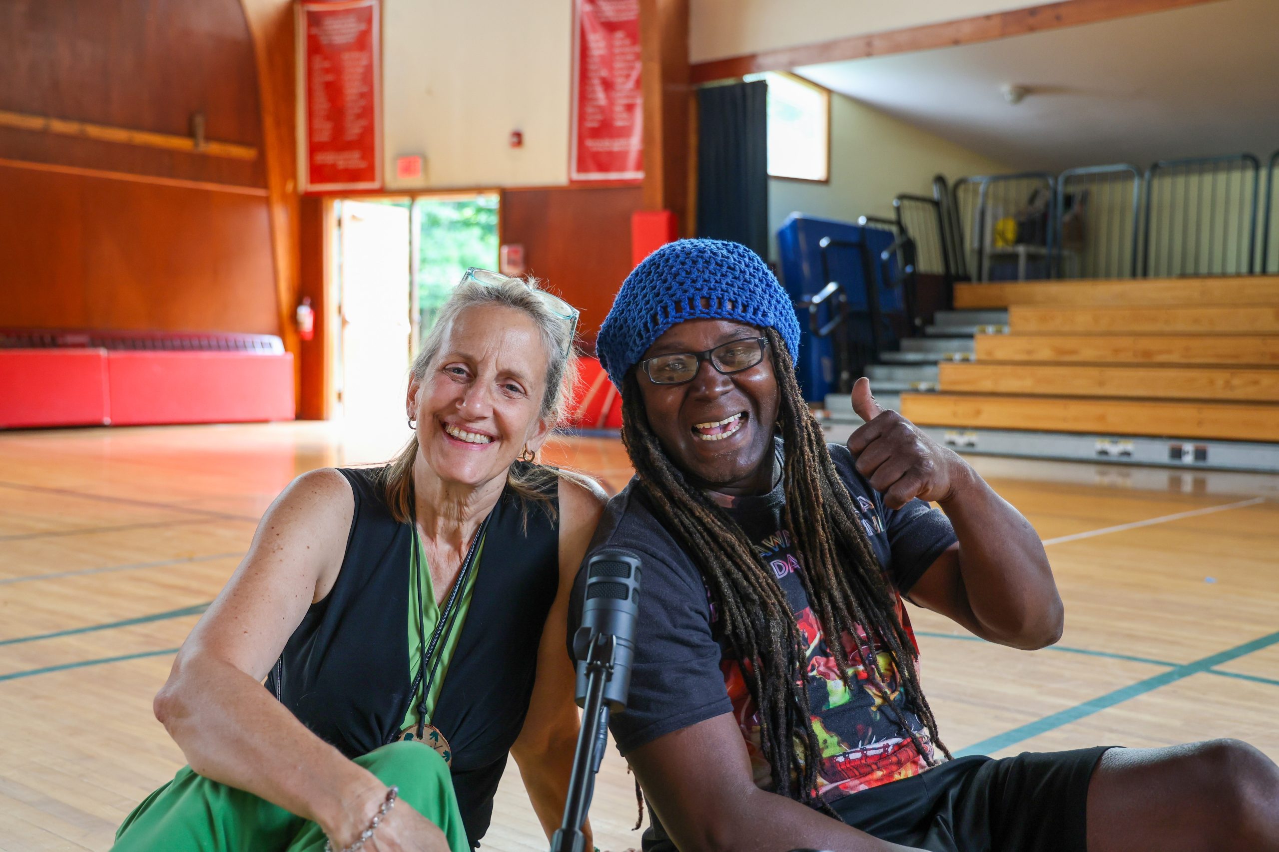 2 Adults sitting in front of a podcast microphone smiling and giving a thumbs up, inside a gymnasium with open doors and sunlight shining inside
