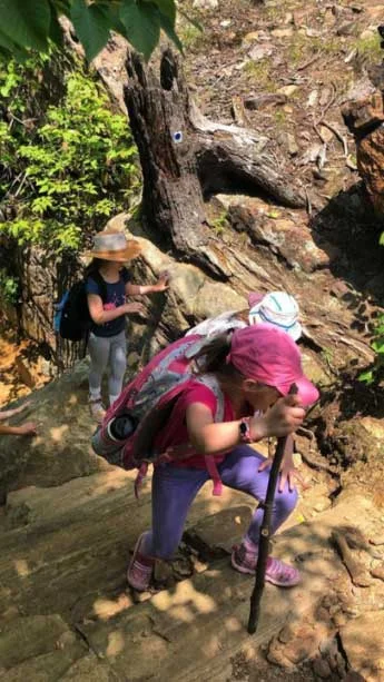 Three campers hiking up a rocky trail in a forest, one wearing a pink cap and carrying a walking stick, and the other in a wide-brimmed hat, and the third in a white bucket hat.