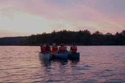 A group of campers in life jackets paddling together in a canoe on a lake at sunset. The sky is painted with soft pink and orange hues.
